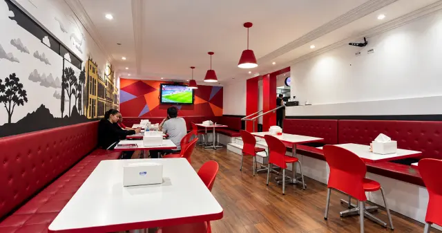 Vista do salão do City Burger com sofás vermelhos, mesas brancas, luminárias pendentes e televisão ao fundo